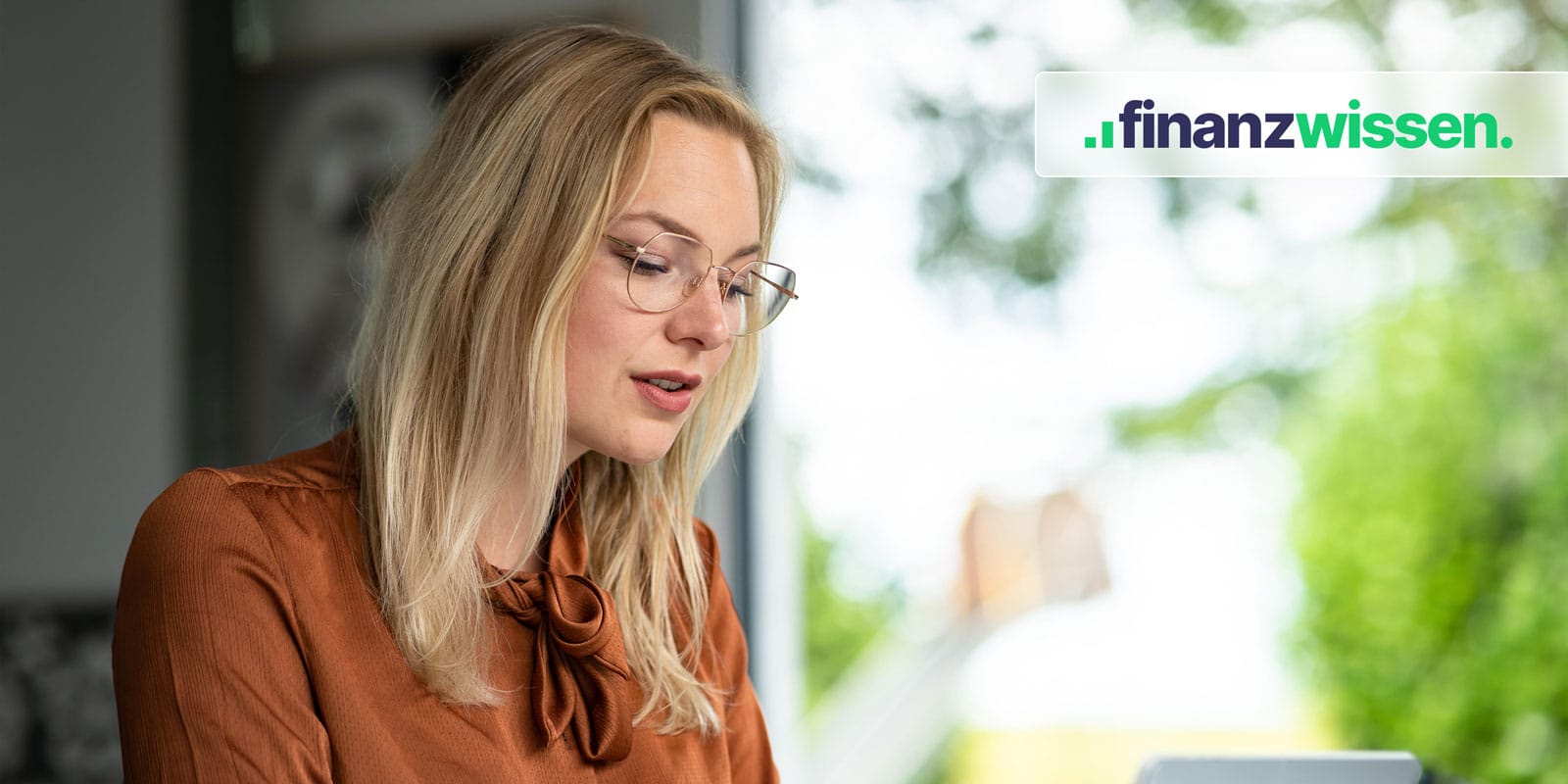 Blonde Frau mit Brille arbeitet am Laptop. Im Hintergrund ein Fenster mit Blick ins Grüne.